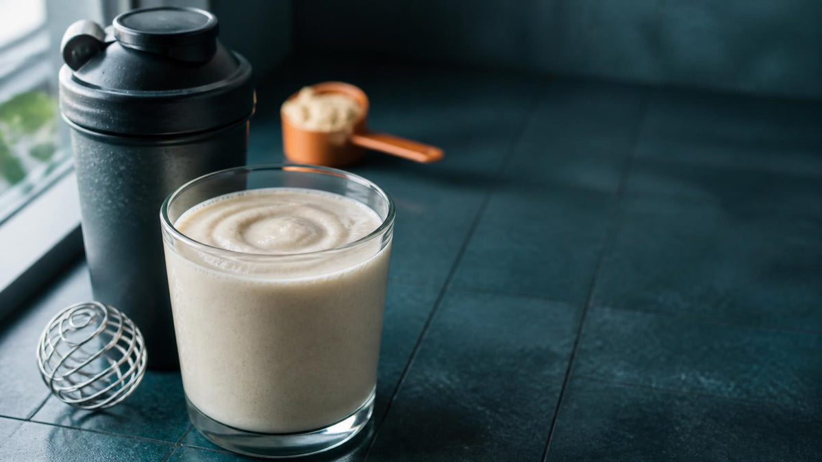 Glass of smooth vanilla protein shake beside a dark shaker bottle with a whisk ball on a teal counter