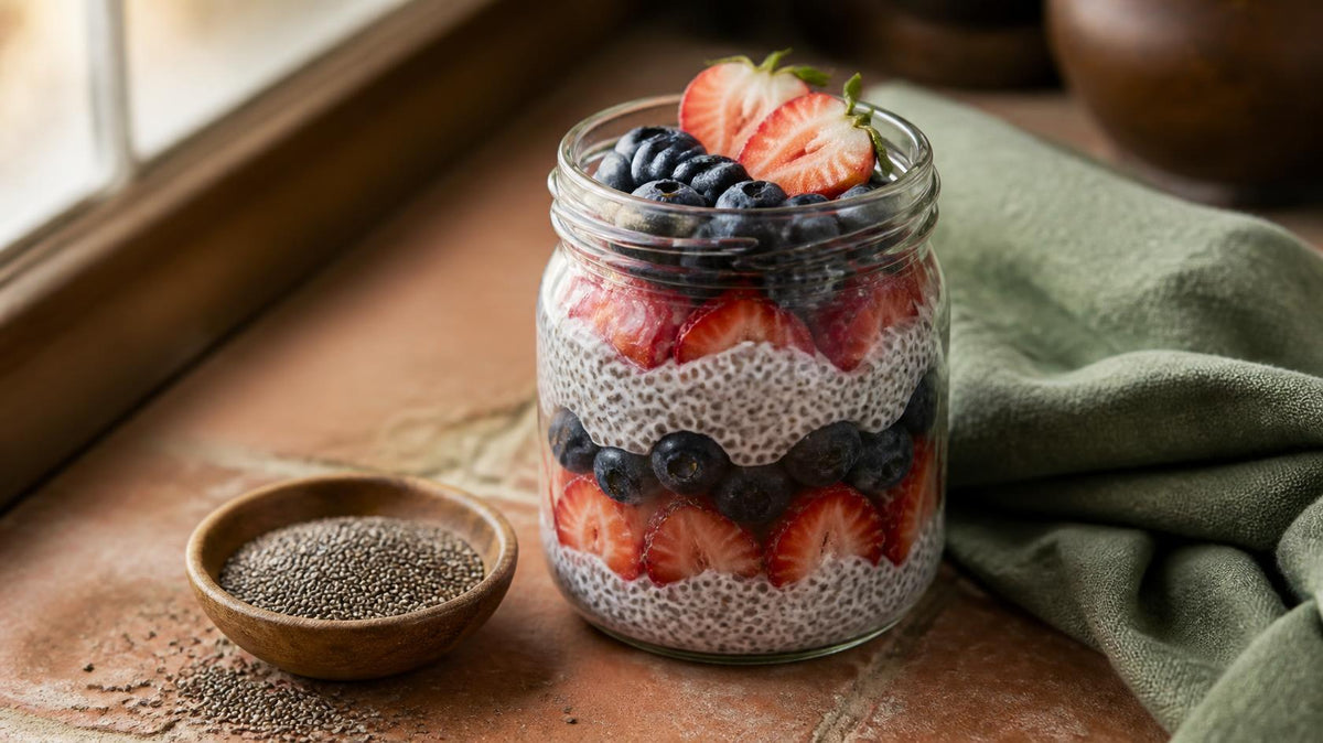 Glass jar of chia pudding layered with strawberries and blueberries on a terracotta tile beside a wooden bowl of chia seeds