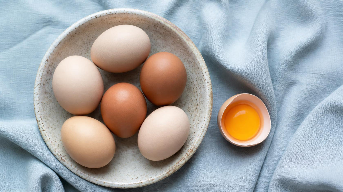 Brown and cream eggs in a speckled ceramic dish on pale blue linen with one cracked egg showing a bright yolk