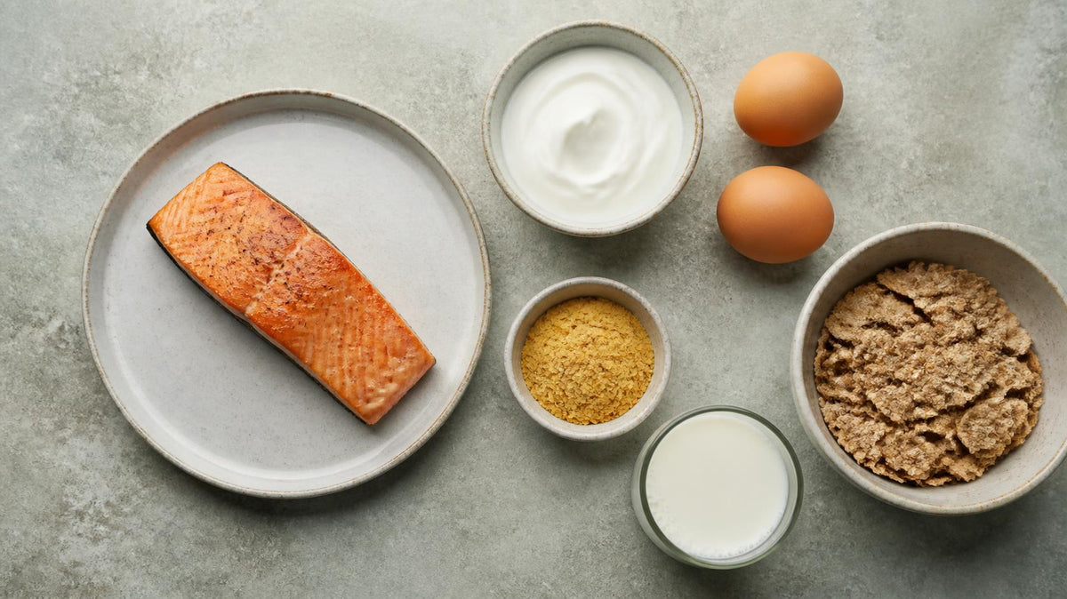 Flat lay of cooked salmon, yogurt, brown eggs, nutritional yeast, milk and cereal on a pale grey-green concrete surface