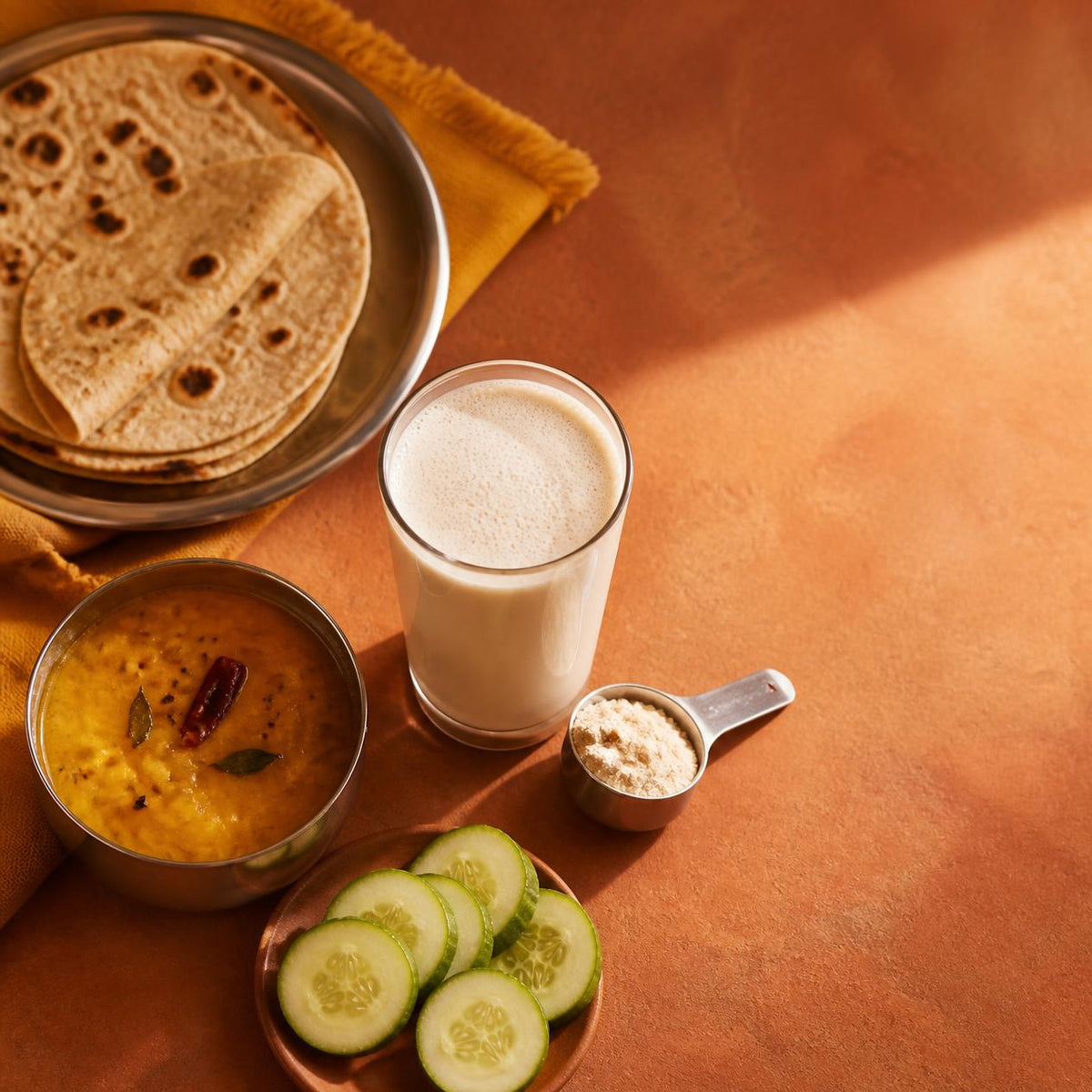Indian meal of dal, rotis and cucumber slices beside a glass of whey protein shake and a steel scoop